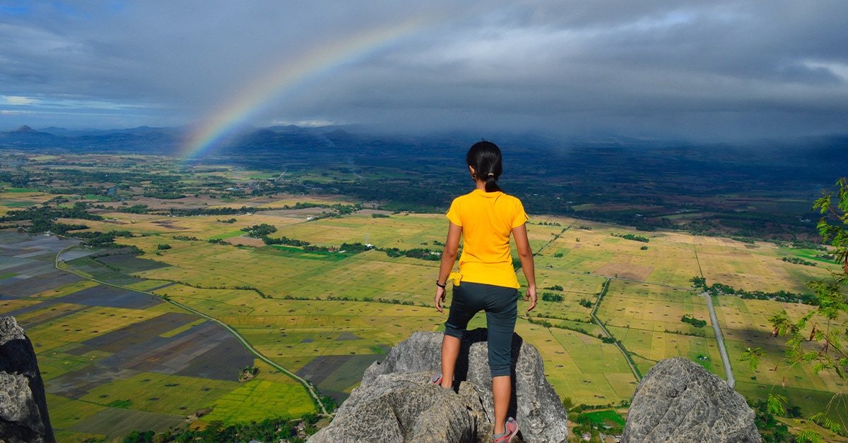 person in yellow shirt looking at rainbow from atop rocky outcropping on hillside.